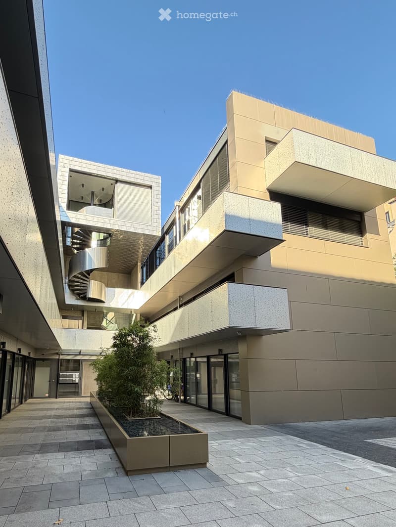 Modern multi-story building with clean lines and contemporary design, featuring a courtyard with a small water feature and greenery.