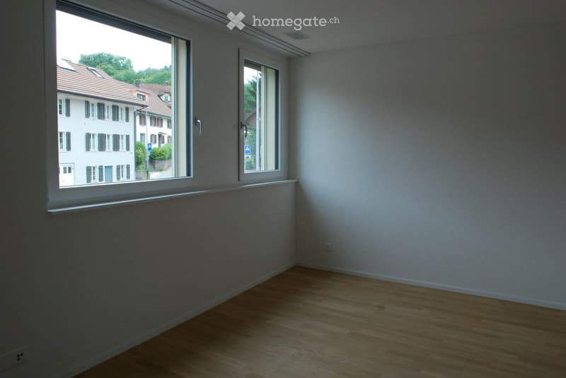 Empty room with wooden flooring, white walls, and large windows offering a view of neighboring buildings and greenery.