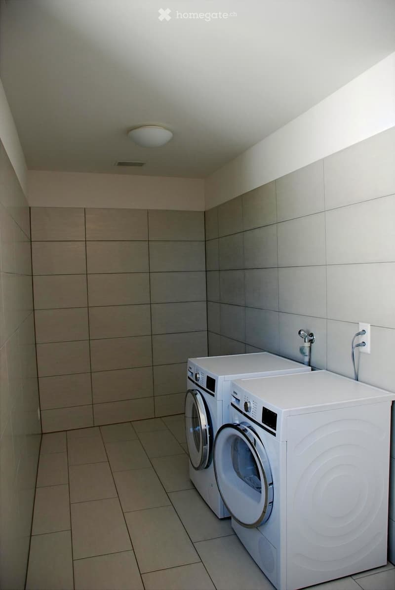 A small, clean utility room with tiled walls and floor, featuring a washing machine and dryer side by side.