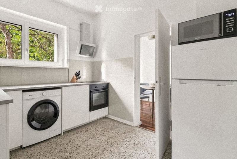 Modern kitchen with white cabinets, stainless steel appliances, and a small dining area visible through an open doorway.