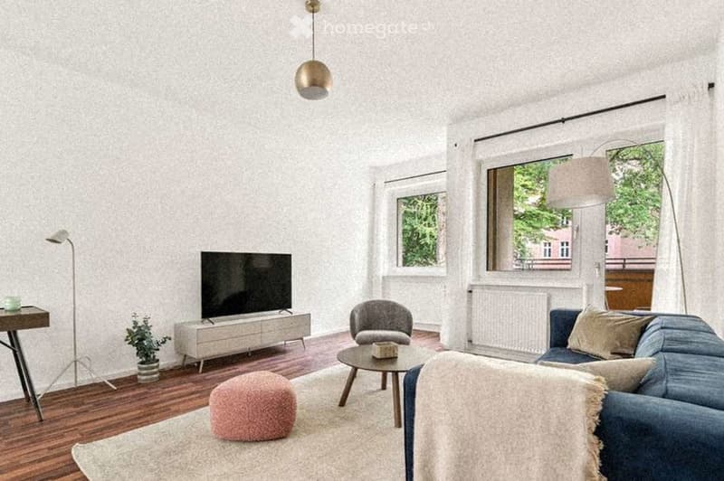 Modern living room with light-colored walls, wooden flooring, and large windows. Features a blue sofa, grey armchair, pink ottoman, coffee table, TV stand, and a small table with a potted plant.