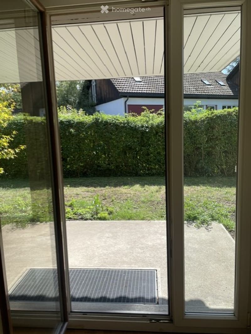 View through a large window showing a well-maintained garden with a concrete patio, neatly trimmed hedges, and neighboring houses visible in the background.