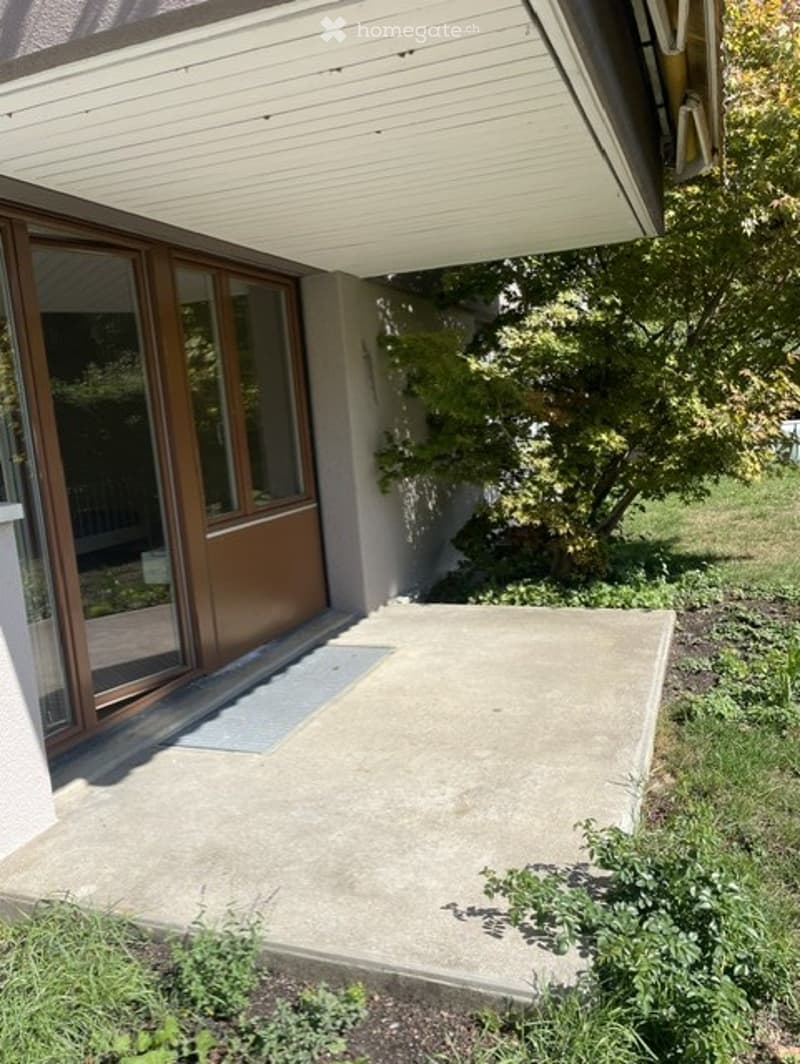 A modern balcony or terrace area with a sliding glass door, concrete floor, and surrounding greenery.