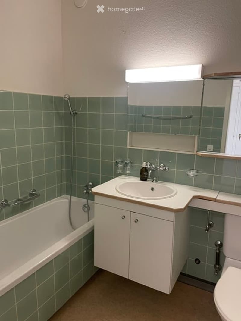 Bathroom with green tiled walls, a white bathtub, a sink with a cabinet, a mirror, and a showerhead.