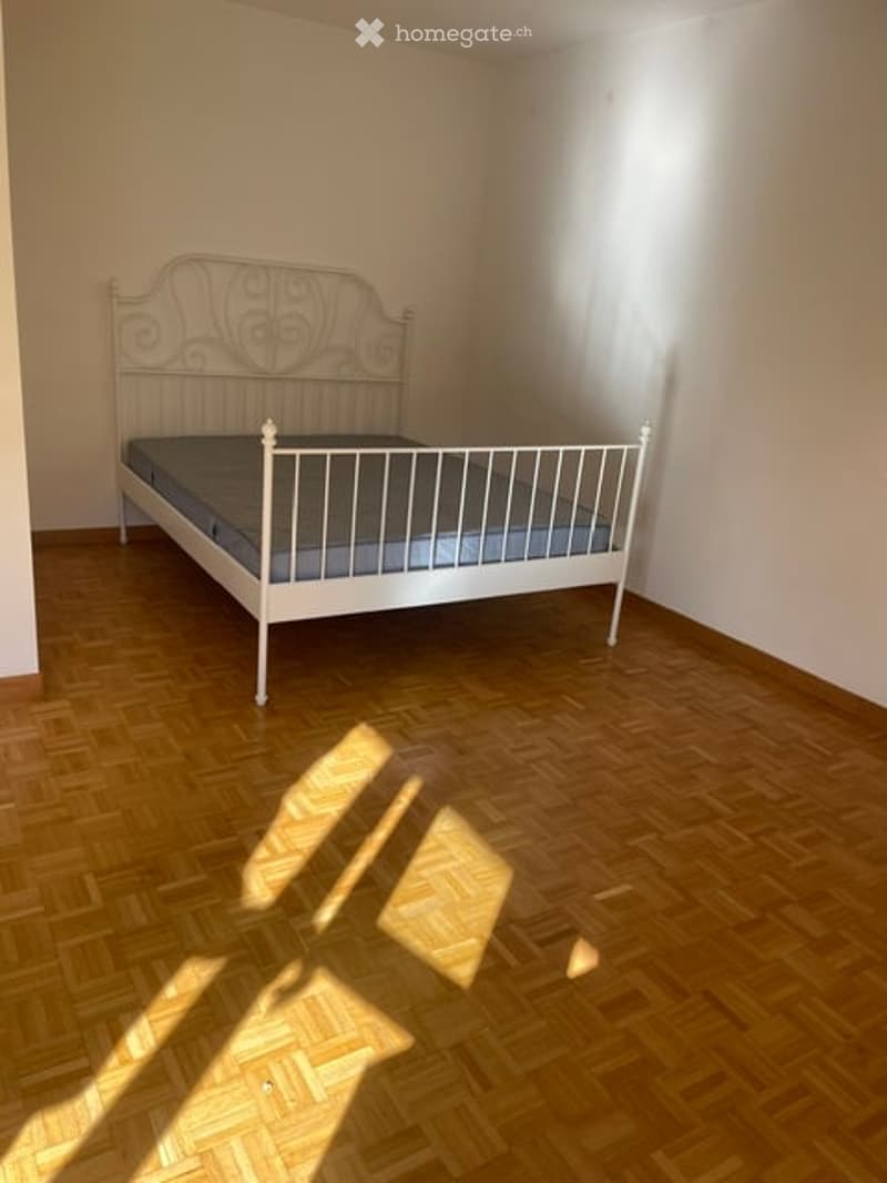 A simple bedroom with a white metal bed frame and blue mattress, set on a wooden floor with natural light coming through a window.