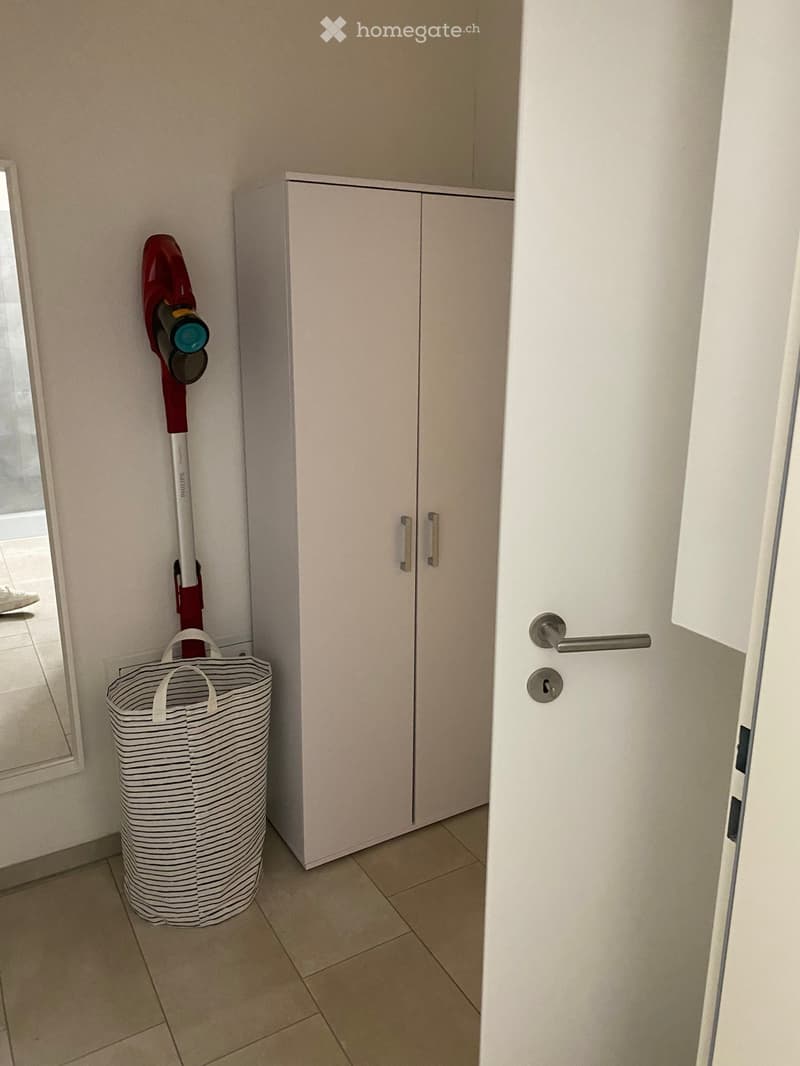 A narrow hallway with a white cabinet, a vacuum cleaner hanging on the wall, and a striped laundry basket on the floor.