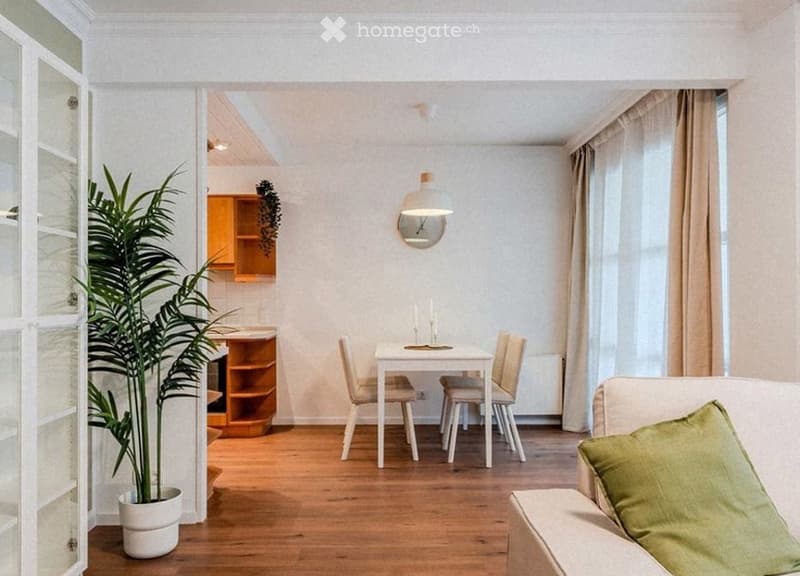 A modern, open-concept living and dining area with light wooden flooring, white walls, and large windows with beige curtains. Features include a white sofa with green cushions, a small dining table with four chairs, a potted plant, a wooden cabinet, and a