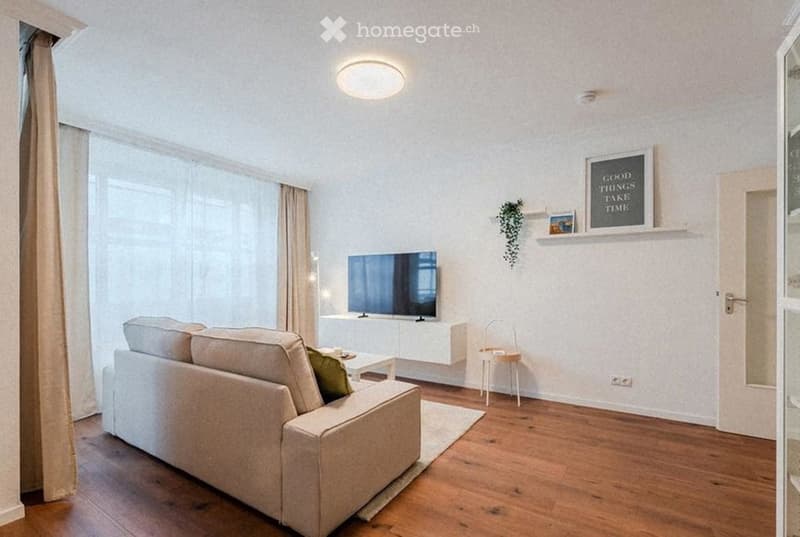 Modern living room with light-colored sofa, coffee table, television, and a small chair near the window. The room features wooden flooring, large windows with curtains, and a decorative shelf with a picture frame and a small plant.