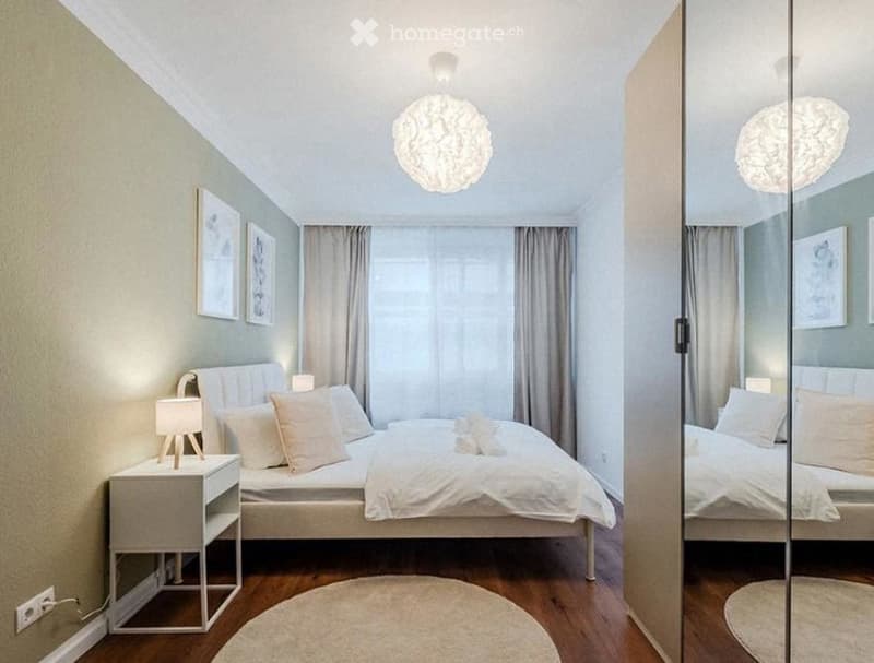 Modern bedroom with a large bed, nightstand, lamp, and mirror. The room features light-colored walls, wooden flooring, and decorative elements like picture frames and a round rug.