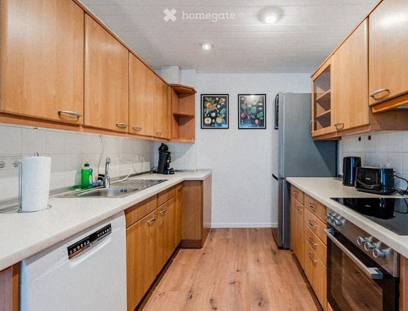 Modern kitchen with light wooden cabinets, stainless steel appliances, and white countertops. Features include a refrigerator, dishwasher, sink, coffee maker, toaster, and oven. Two framed artworks adorn the wall.
