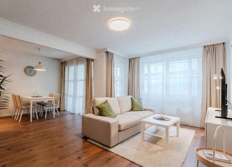 Modern living room with a light-colored sofa, coffee table, dining area with a table and chairs, and a television stand. The room features large windows with curtains, wooden flooring, and a small plant in the corner.