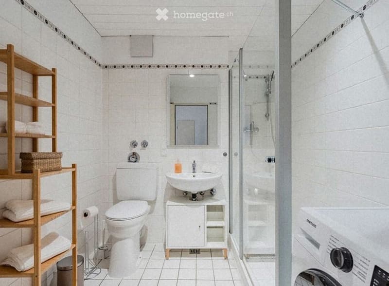 Modern bathroom with white tiles, a toilet, sink, mirror, shower, wooden shelf with towels, and a washing machine.