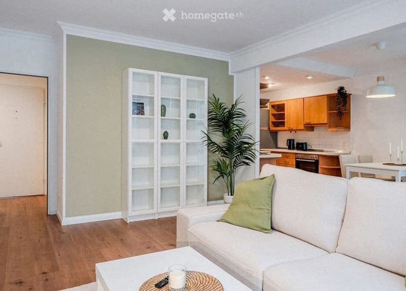 Bright living room with a white sofa, coffee table, bookshelf, and a potted plant. Adjacent kitchen area with wooden cabinets and a dining table.