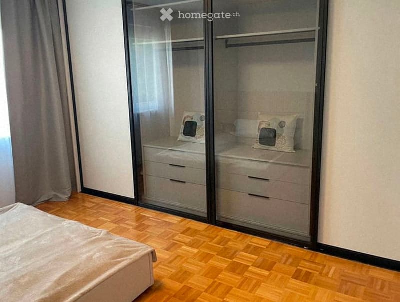 Modern bedroom with wooden flooring, a large mirrored wardrobe, and a neatly made bed with pillows.
