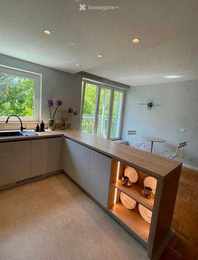 Modern kitchen with light grey cabinets, a wooden countertop, built-in shelving with plates and cups, a sink with a faucet, and a small dining area with a table and chairs. The room features large windows with a view of greenery outside.