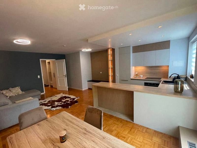 Open-plan living and dining area with a grey sofa, coffee table, dining table with chairs, and a modern kitchen island with cabinets, sink, and stove. The space features wooden flooring, recessed lighting, and a cowhide rug.