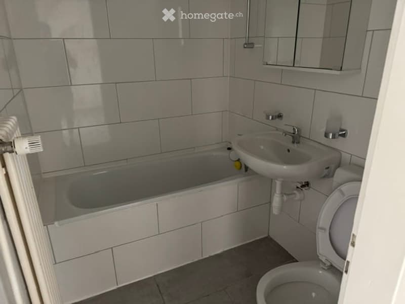 A small bathroom with white tiled walls and floor, featuring a bathtub, sink, toilet, and a mirror above the sink. A radiator is visible on the left side.