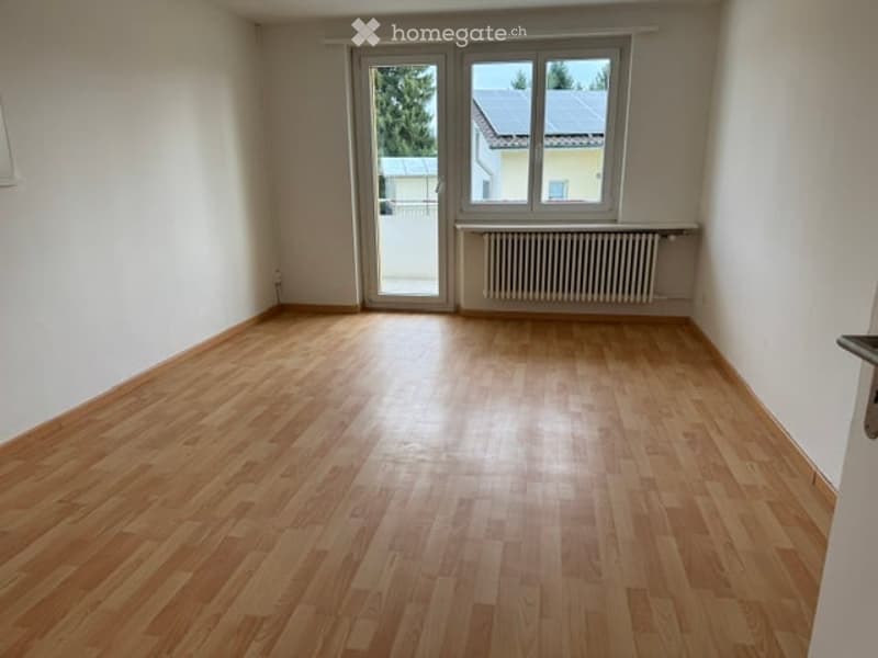 Empty room with wooden flooring, white walls, and a large window with a radiator below it. The room has a sliding glass door leading to an outdoor area.