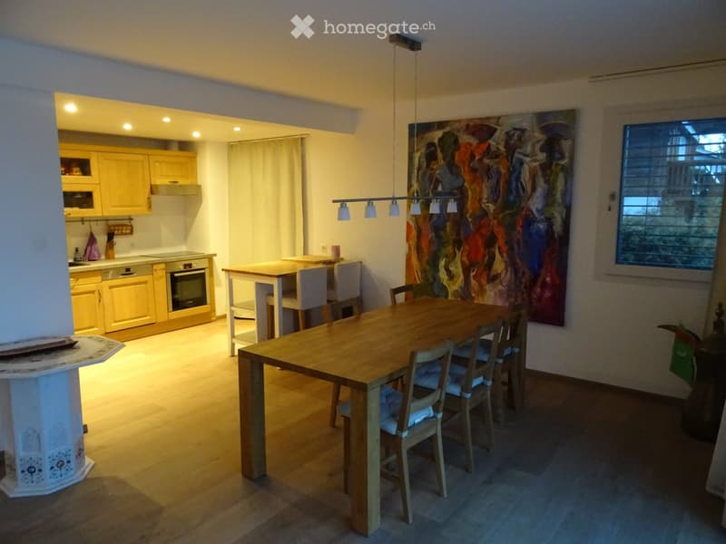 Open-plan living and dining area with a wooden dining table and chairs, a kitchen with cabinets, an oven, and a dishwasher, illuminated by pendant lights and warm lighting.