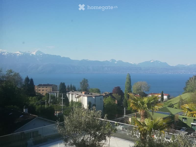 Panoramic view of a lake and mountains from a rooftop terrace, with surrounding buildings and greenery.