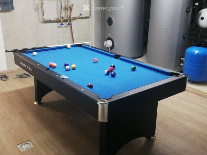 A room with a pool table and scattered pool balls, featuring a wooden floor and visible pipes and equipment in the background.