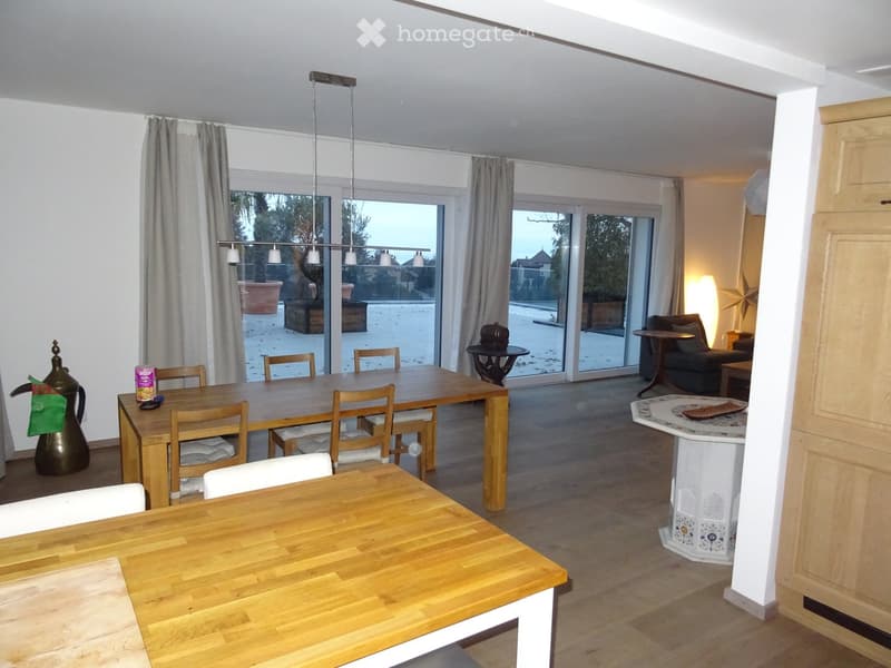 Bright, open-plan living and dining area with wooden flooring, large windows, and a view of a snowy outdoor space. Includes a dining table with chairs, a sofa, a small side table, and decorative items.
