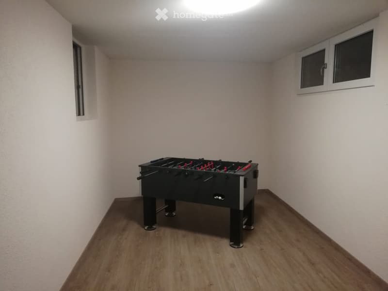 A small, unfurnished room with wooden flooring and white walls, featuring a foosball table as the sole piece of furniture.