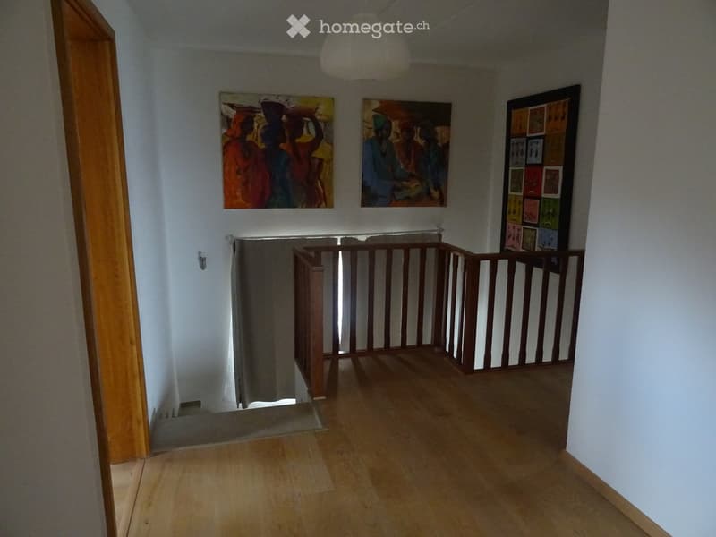 Spacious hallway with wooden flooring, white walls, and a wooden railing leading to an upper level. Three paintings are displayed on the walls, and a pendant light hangs from the ceiling.