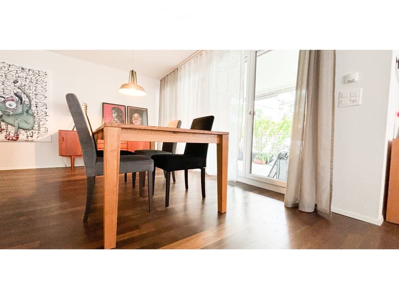 Dining room with wooden table, chairs, pendant light, and artwork on the wall. Large window with curtains leads to a balcony.
