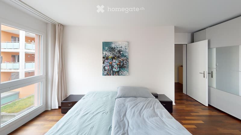 Modern bedroom with a single bed, nightstands, a colorful abstract painting on the wall, and a large window with a view of a balcony and greenery.