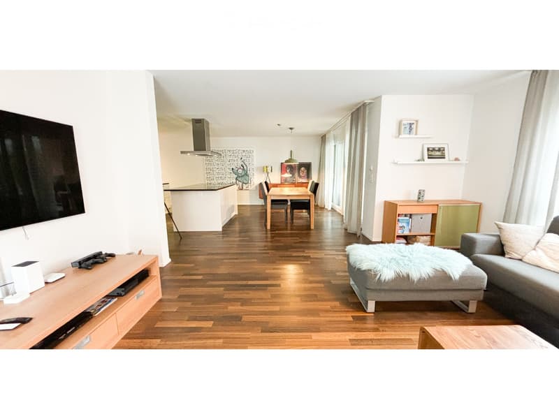 Modern open-plan living and dining area with wooden flooring, a grey sofa with an ottoman, a coffee table, a TV stand, a kitchen island, a dining table with chairs, and built-in cabinets and shelves.