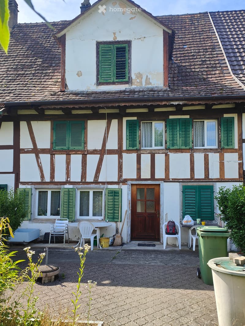 Two-story house with white and brown exterior, green shutters, and a wooden front door. Outdoor area includes a table, chairs, and a backpack.