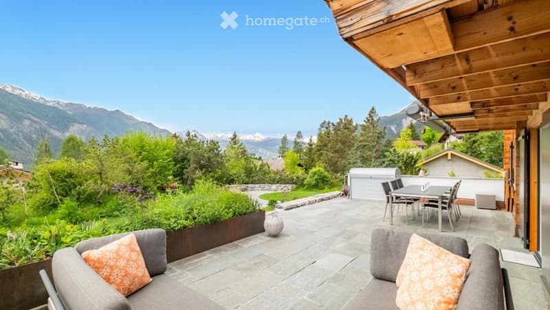 Outdoor patio with stone tiles, two grey sofas with orange cushions, a dining table with chairs, and a scenic mountain view.