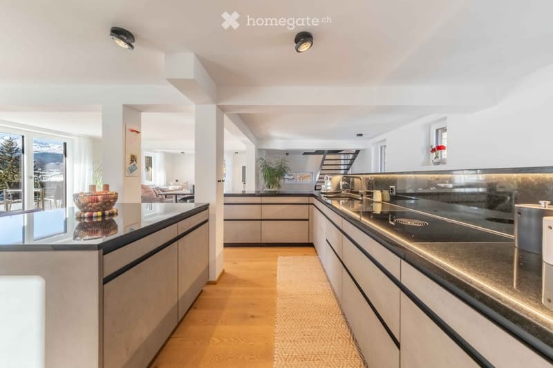 Modern kitchen with white cabinets, dark countertops, and stainless steel sink. Features include recessed lighting, a bowl on the counter, and a view of a living area with seating.