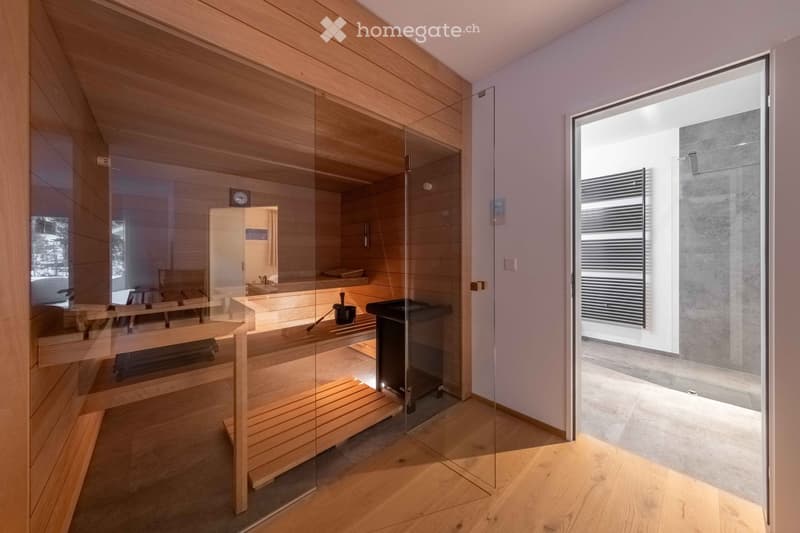 Modern bathroom with wooden sauna bench, black bucket, and black stove, featuring glass walls and wooden flooring.