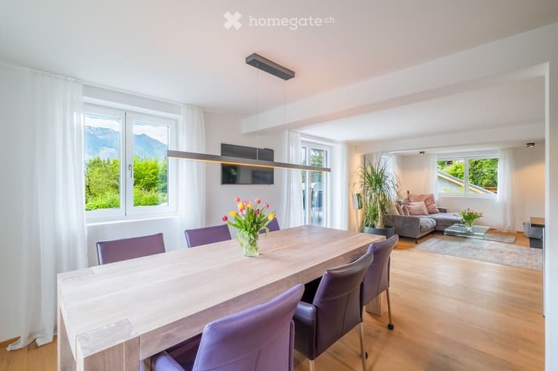 Open-plan living and dining area with modern furniture, large windows offering a scenic mountain view, a dining table with chairs, a sofa, and decorative plants.