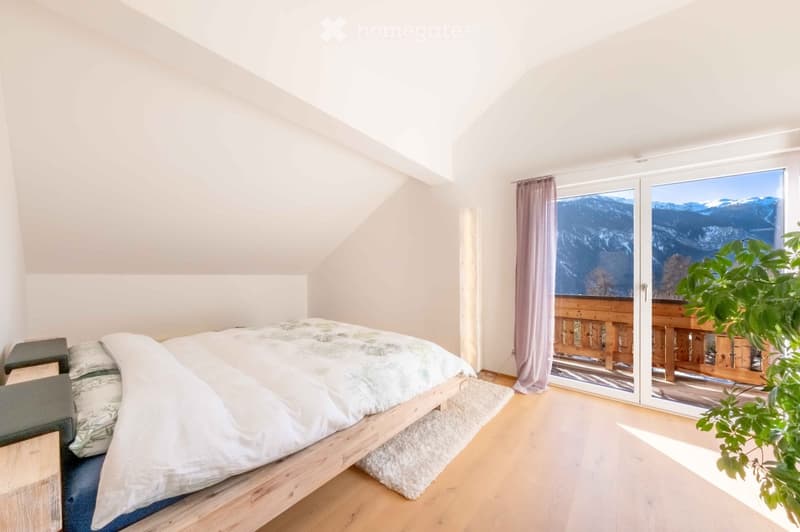 Bright bedroom with wooden flooring, a large bed with white bedding, a nightstand, a chair, and a plant near a large window offering a scenic mountain view.