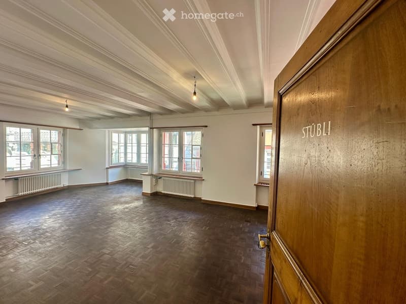 Empty room with wooden flooring, white walls, large windows, radiators, and exposed wooden beams on the ceiling.