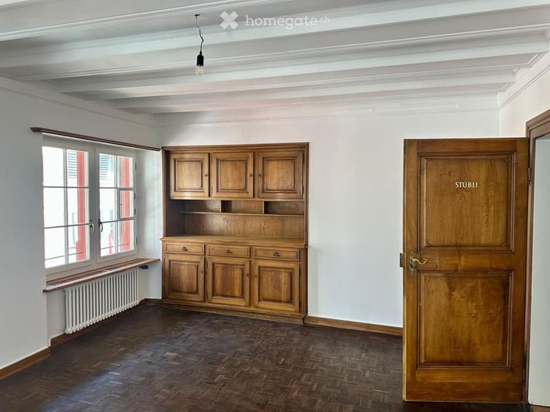 Empty room with wooden floor, white walls, large window with radiator, wooden cabinet, and wooden door labeled "STÜBLI".