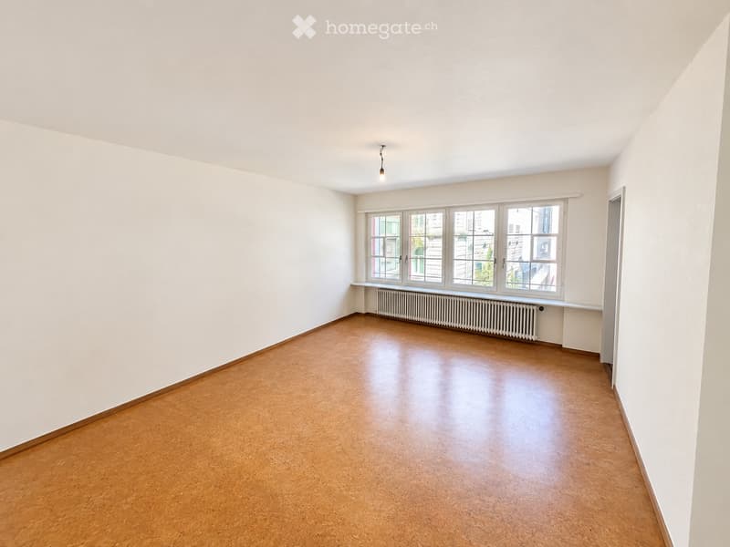 Empty room with light-colored walls, wooden flooring, and large windows with a radiator below. No furniture or personal items are present.