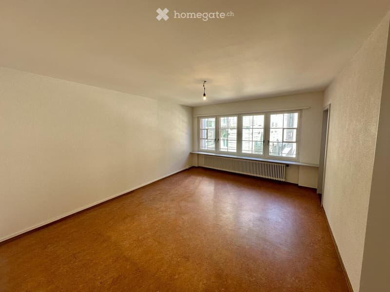 Empty room with wooden flooring, white walls, large windows, and a radiator under the windows.