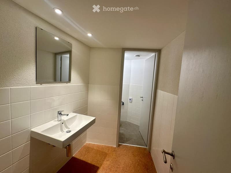 Modern bathroom with a white sink, chrome faucet, and a large mirror above it. The walls are tiled in white and beige, and there is a doorway leading to a shower area.