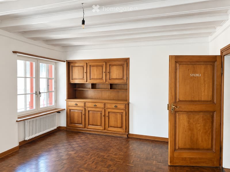 Empty room with wooden floor, white walls, large window, wooden cabinet, and a wooden door.