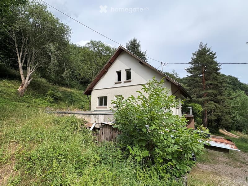 A single-story house with a pitched roof, surrounded by greenery and trees. The house has multiple windows and a small balcony on one side.
