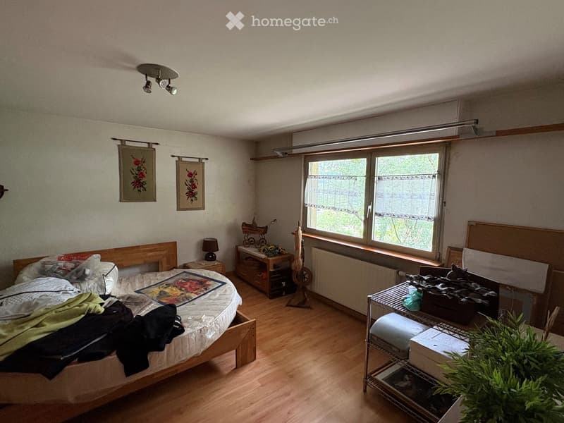 A well-lit bedroom with wooden flooring, a large bed with a colorful quilt, a nightstand with a lamp, a small table with a toy, and a window with blinds. There is also a radiator under the window and a plant on a metal shelf.