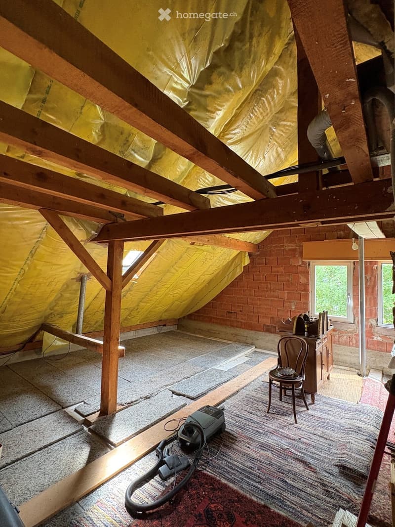 An attic space with exposed wooden beams, insulation, and a few pieces of furniture including a chair, a small cabinet, and a vacuum cleaner on the floor.