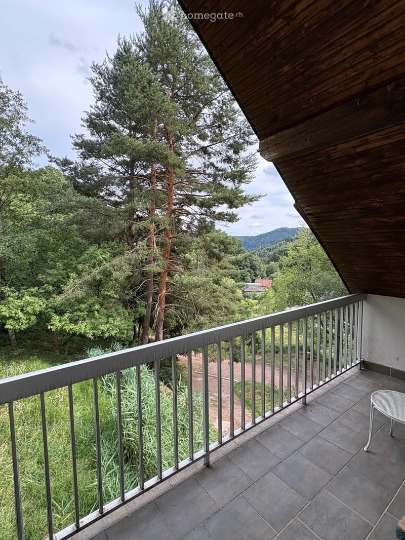 Balcony with metal railing, tiled floor, and a white chair, overlooking a forested area with trees and hills.