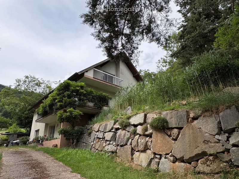 Two-story house with a balcony, surrounded by greenery and a stone retaining wall.
