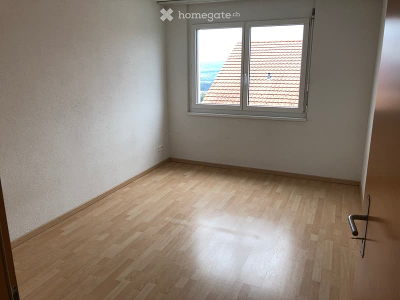 Empty room with light wooden flooring and white walls, featuring a large window with a view of a tiled roof.