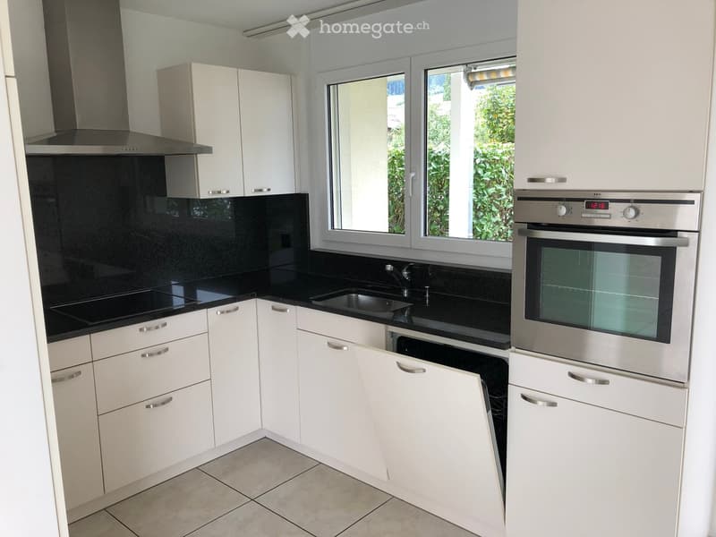 Modern kitchen with white cabinets, black countertops, stainless steel appliances, and a large window overlooking greenery.